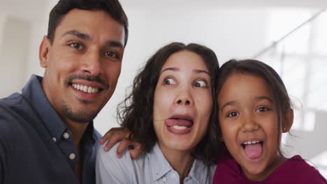 Glückliche-Hispanische-Eltern-Und-Tochter-Umarmen-Sich-Und-Machen-Im-Wohnzimmer-Lustige-Gesichter
