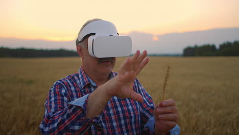 Senior-adult-farmer-in-a-virtual-reality-helmet-in-a-field-of-grain-crops.-In-the-sunset-light-an-elderly-man-in-a-tractor-driver-uses-virtual-reality-glasses.-VR-technologies-and-modern-agribusiness