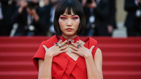 a poised figure in striking red attire, adorned with exquisite jewelry, captures the essence of elegance and style, showcasing bold makeup with a captivating expression amidst a crowd of photographers.