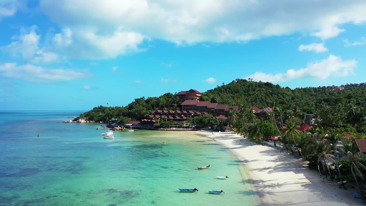 Anchored boats on calm turquoise lagoon in front of white sandy beach with palm trees near holiday resort in Thailand