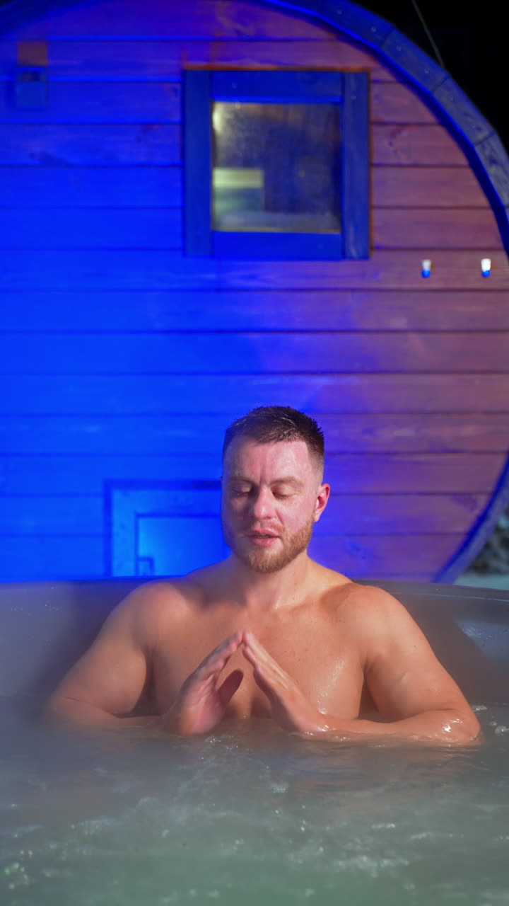 Bearded Caucasian man sits in a hot tub. Man rubs his face and then leans back relaxed in jacuzzi. Vertical video.