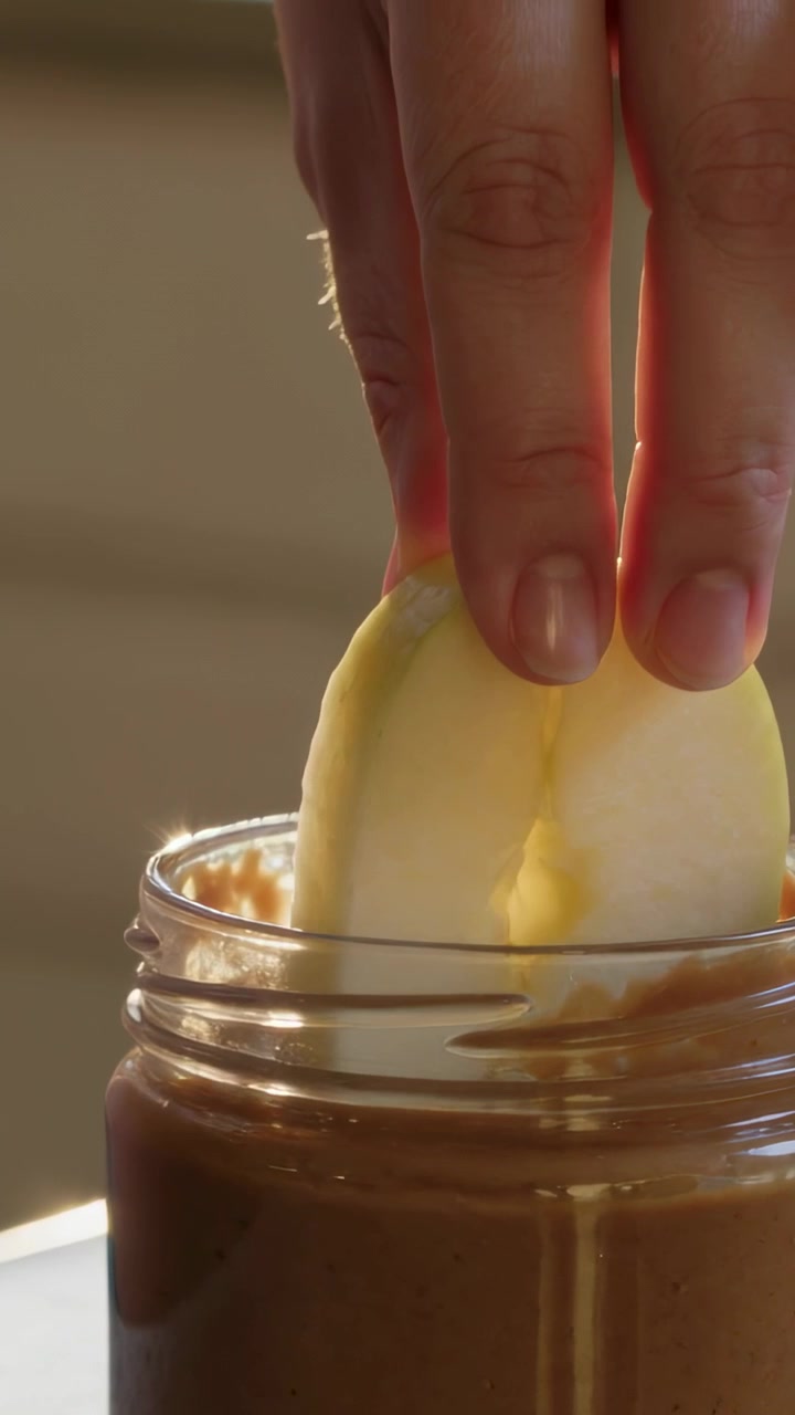 Video verticale: Una mano che intinge una fetta di mela in un barattolo in cucina, ricoprendola con una crema spalmabile marrone.