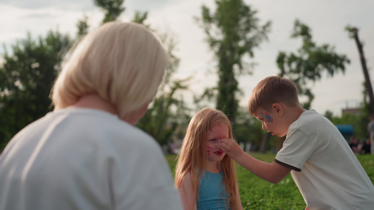 young boy carefully wiping sister cat face paint with soft tissue under warm summer sun in green park while sister sits on grass enjoying tender sibling moment during family picnic