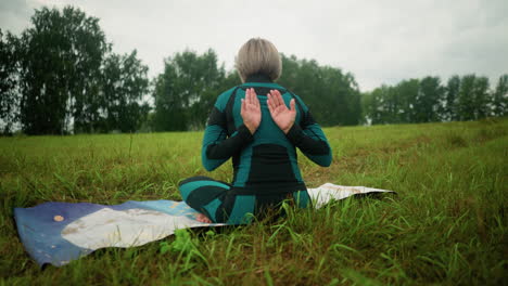 back view of middle-aged woman sitting on mat in sukhasana pose practicing yoga with eyes closed, embracing calmness in a vast grassy field under a cloudy sky, with trees lined up in the background