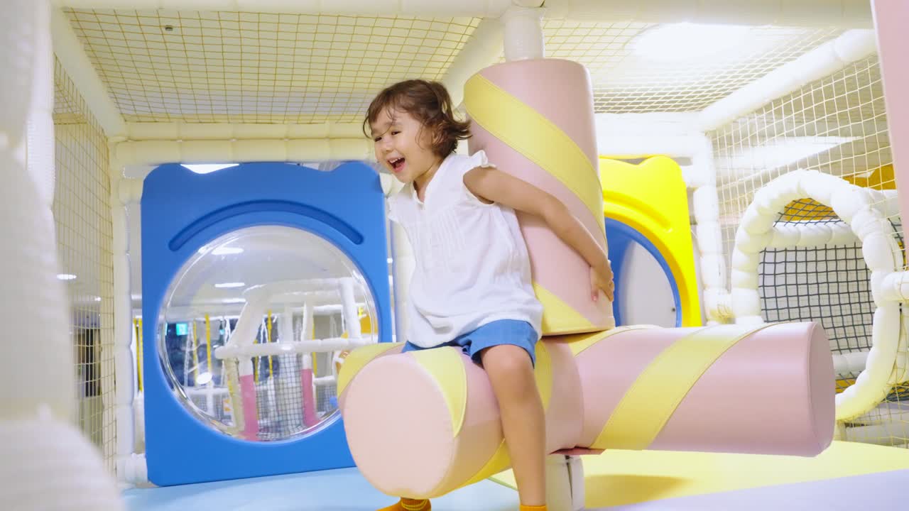 Happy little girl laughs with joy while riding a soft spinning obstacle at a modern indoor playground and entertainment center for kids