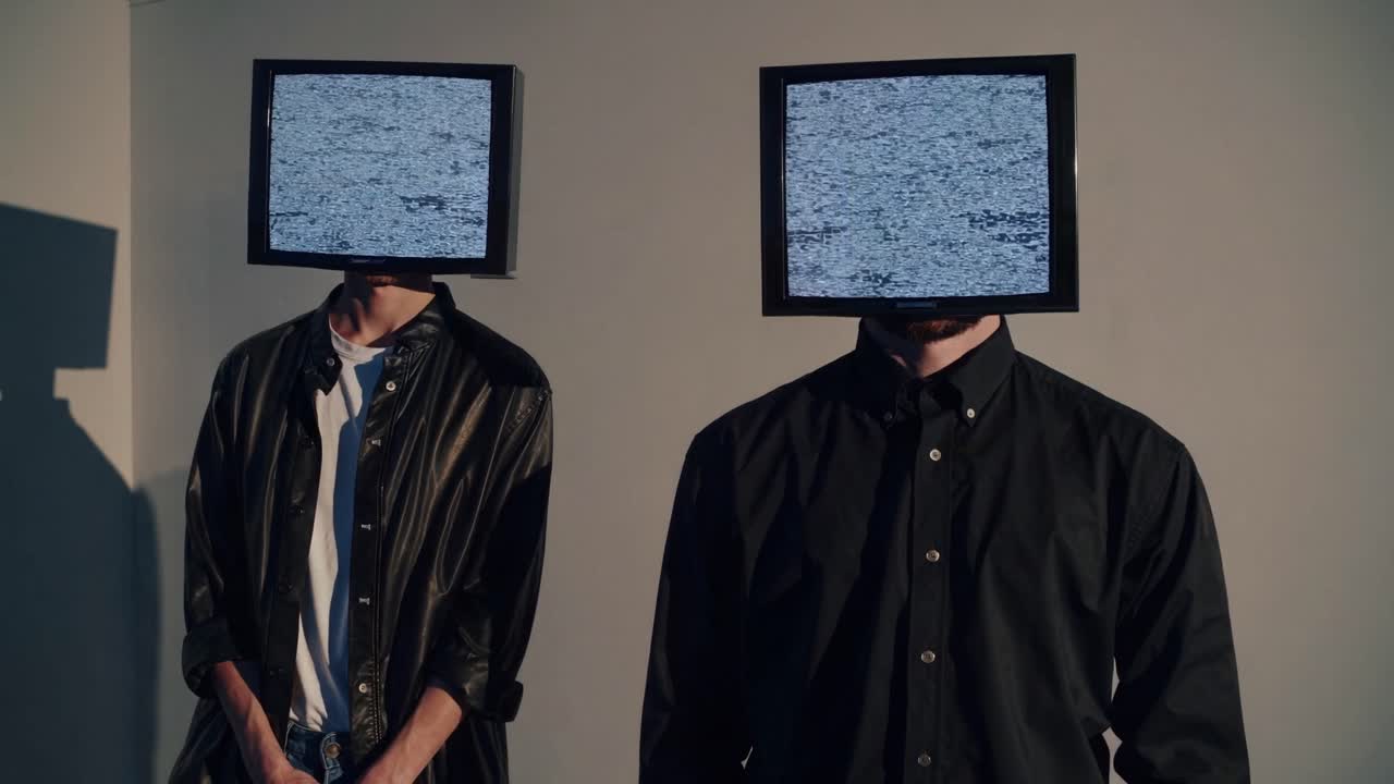 Two figures with TV screens as heads, showing static, stand together in a simple environment, evoking themes of technology and identity in a captivating scene