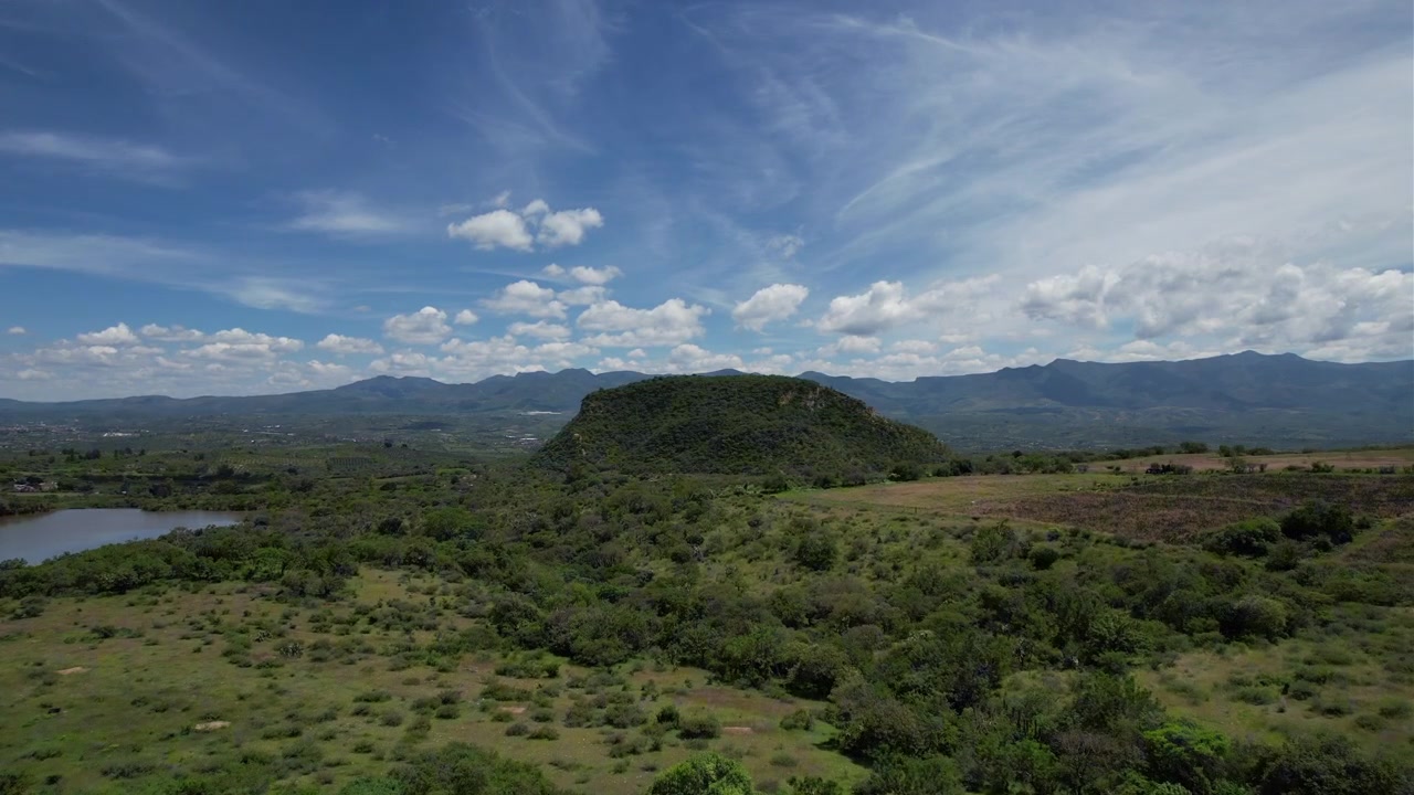 Riprese aeree con drone che si allontanano per rivelare un paesaggio verde e panoramico con una prominente collina dalla cima piatta circondata da campi, vegetazione e catene montuose distanti sotto un cielo blu vibrante.