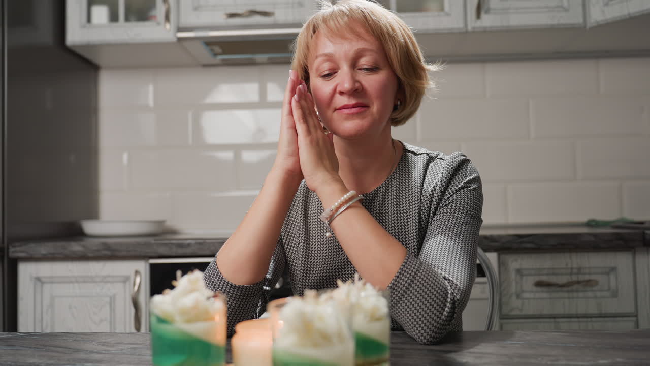 Elegant lady claps hands gently and moves them sideways in joyful admiration of her beautifully crafted candle recipes, set in layered glass cups with green and white wax on tray in kitchen setting
