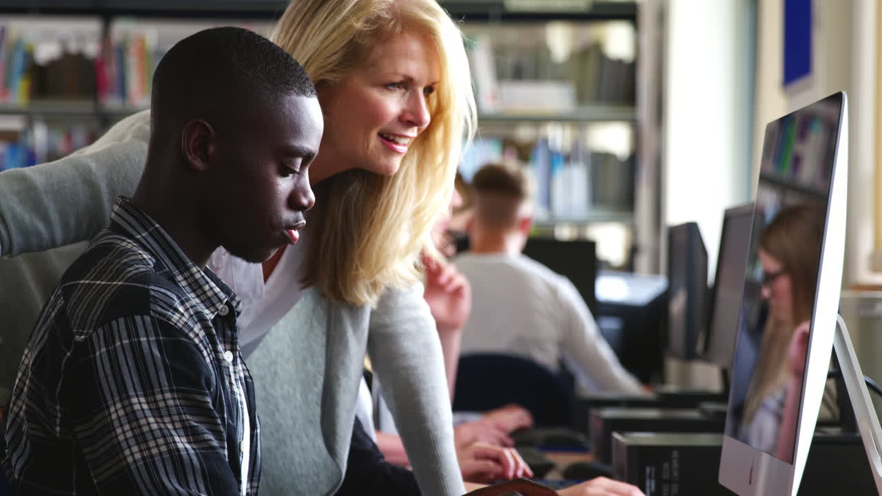 Teacher With Male Student Working On Computer In College Library