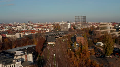 Berlin,-Deutschland-Stadtbild-Mit-Dem-Zug,-Der-In-Eine-Station-Neben-Dem-Berühmten-Gasometer-Und-Dem-Fernsehturm-In-Der-Ferne-Einfährt,-Luftwagen-Herein