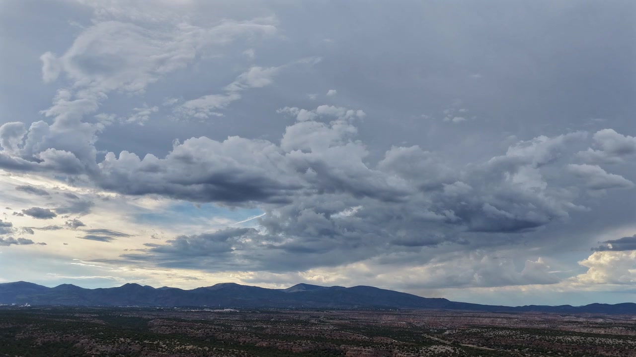 4K drone timelapse of stormy, moody clouds drifting during dusk. The grey overcast sky blankets a vast, rugged mesa landscape and distant mountains across Los Alamos County, New Mexico.