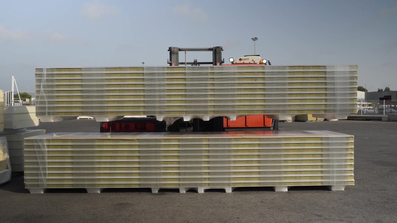 Outdoors during the day, a front view of a stack of insulation panels, a forklift truck lifting one of the two stacks