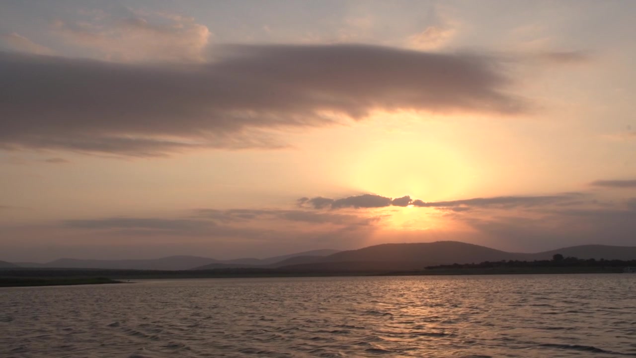 Vue panoramique spectaculaire d'un coucher de soleil doré éclatant sur un grand lac aux ondulations, avec des rayons de soleil perçant les nuages derrière une magnifique silhouette de crêtes montagneuses ondulantes en Afrique du Sud