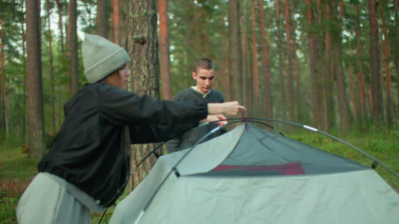 lady secures red rope on tent pole during forest camping while her friend stands nearby watching with hand on waist, surrounded by tall pine trees and soft outdoor lighting