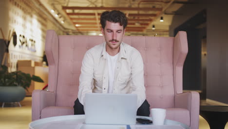 hombre blanco millennial creativo sentado en un sofá usando una computadora portátil en un vestíbulo de la oficina