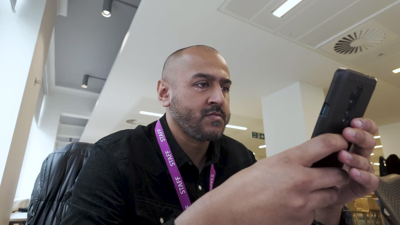 hombre indio asiático con camisa negra usando teléfono inteligente en el interior, expresión enfocada, entorno de oficina