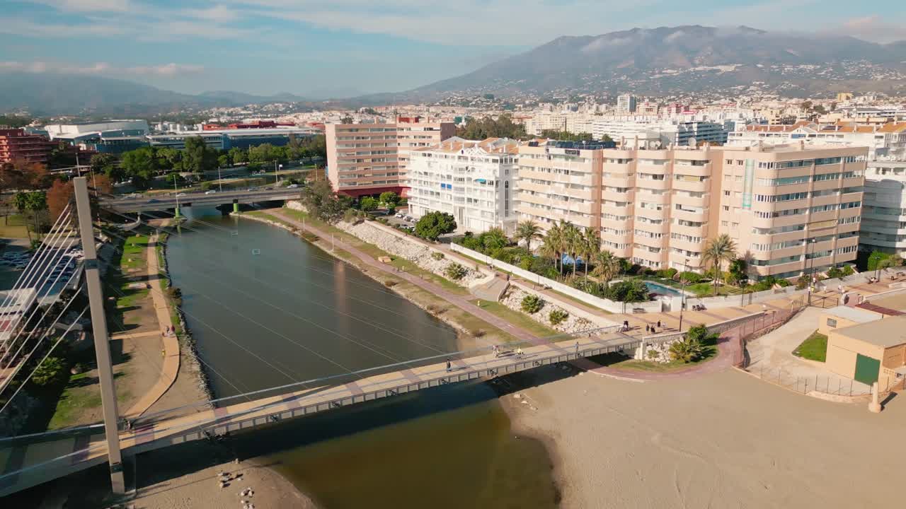 Veduta aerea di Fuengirola, Spagna, che evidenzia il ponte panoramico con persone che camminano, la foce del fiume, gli edifici costieri e lo skyline della città lungo la costa.