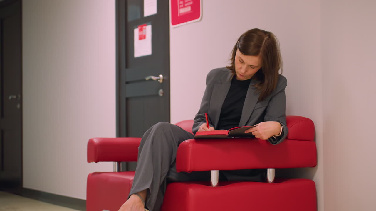 Businesswoman sitting on red chair in office hallway, writing in notebook with red pen, focused on task, preparing for meeting, professional office environment, calm, organized workspace
