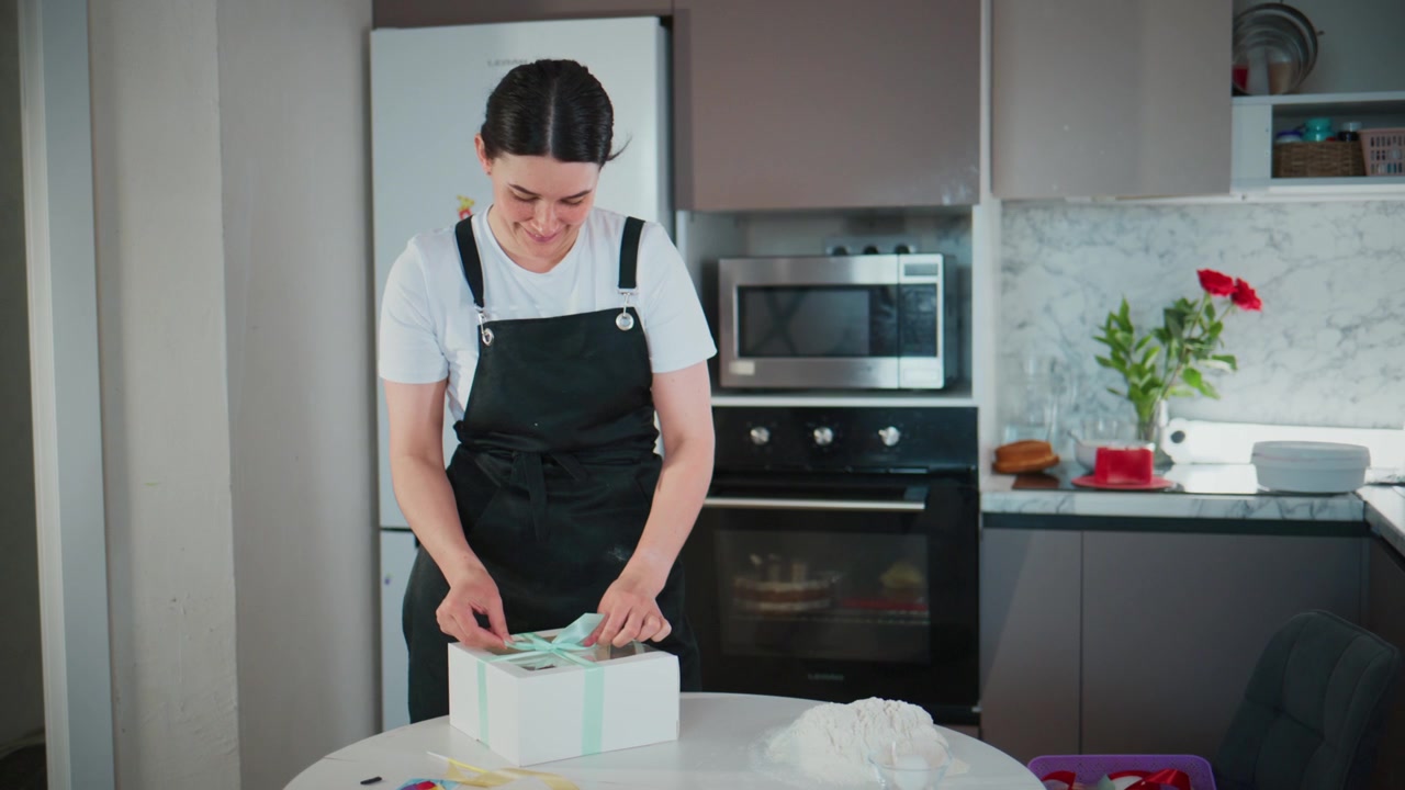 Una donna in cucina incarta con cura una scatola regalo. Sorride dolcemente, concentrata sul suo compito. L'ambiente è moderno, pulito e ben illuminato. Ideale per contenuti relativi a festività, compleanni o regali.