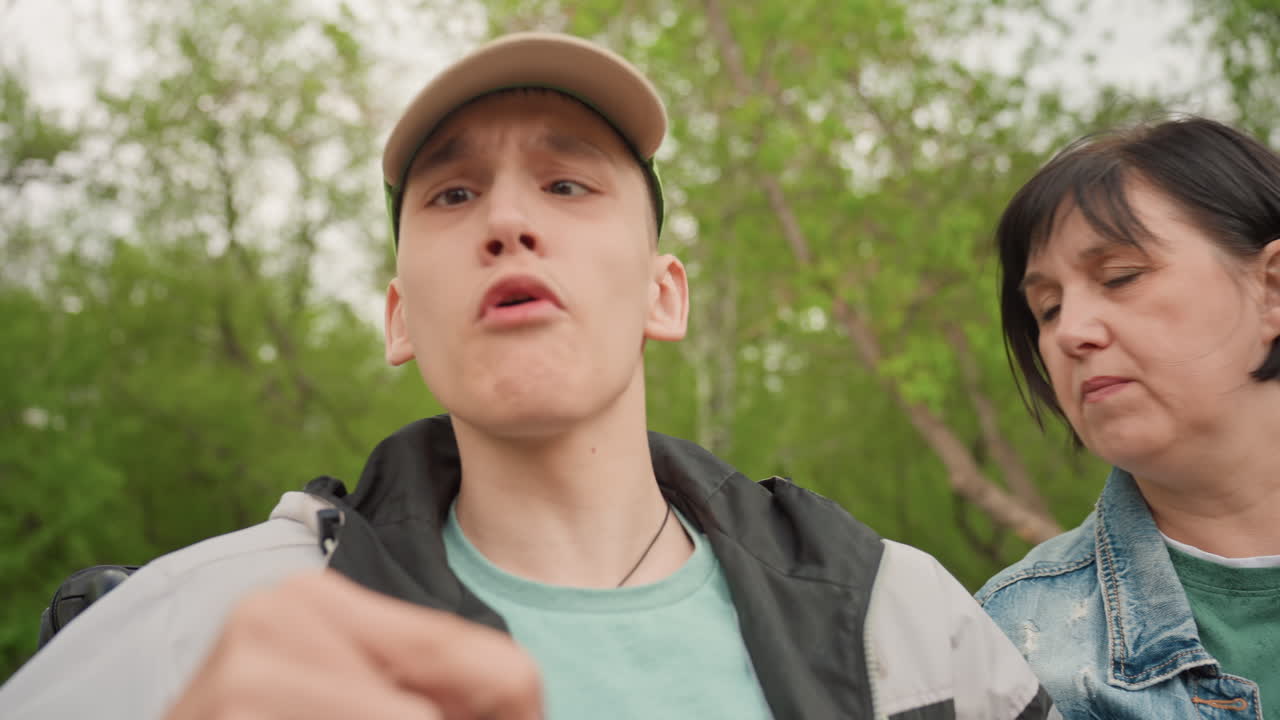 White Blind Young Man Discovering Wallet With Female Caregiver Beside Him, Animated Gestures And Surprised Expression, Outdoor Park Setting, Denim Jacket And Cap, Hands Exploring Object, Candid