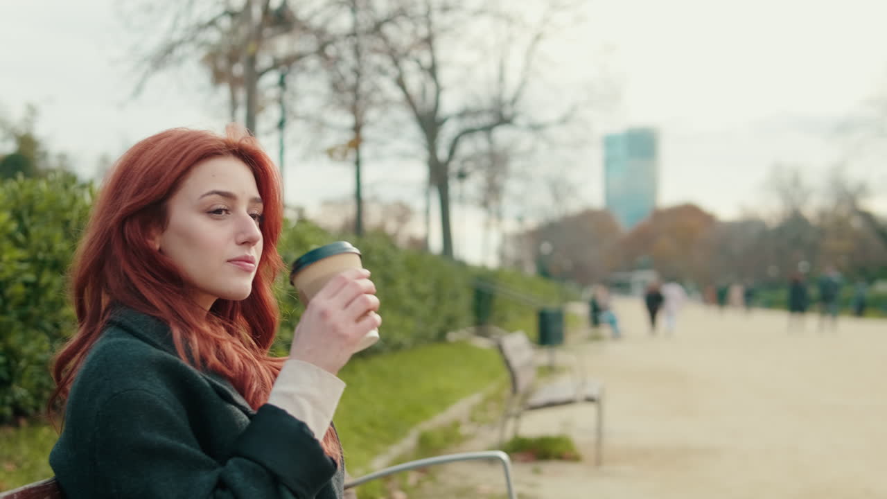 Ung rødhåret kvinde drikker kaffe i en bypark