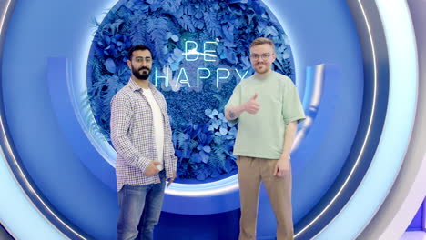two men giving thumbs up in front of a "be happy" neon sign