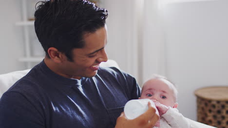 Smiling-Hispanic-father-sits-rocking-his-four-month-old-son,-feeding-him-a-bottle-at-home,-close-up