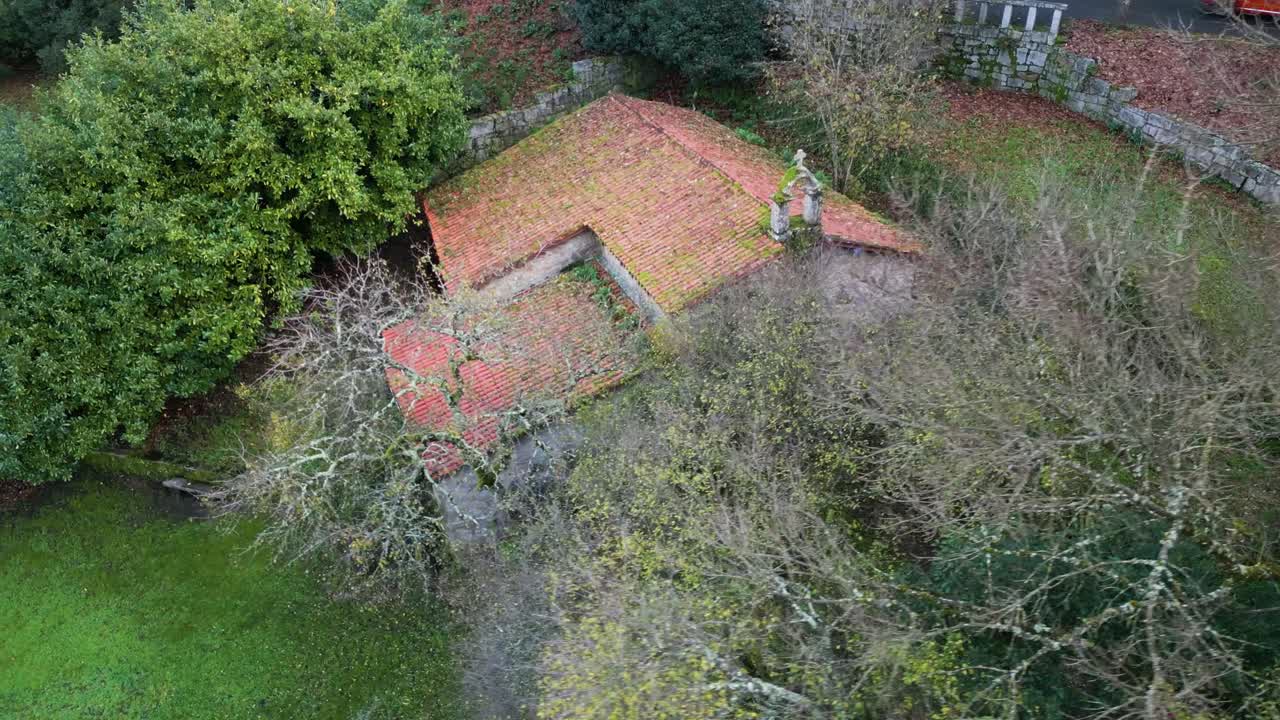 Drone rises above leafless trees to reveal stone cross on mossy roof of Hermitage of Portovello, As Lagoas Ourense Spain