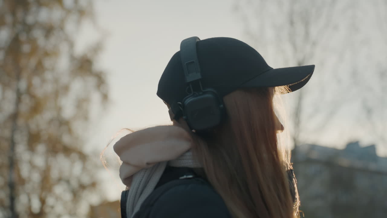 冬のヘッドホンをつけた白人少女。警戒した姿勢で息が白く見え、逆光の都市の建物を背景にキャップとスカーフを着用しています。音楽を聴きながらも、徘徊者やひったくりの可能性を警戒し、周囲を注意深く見回しています。
