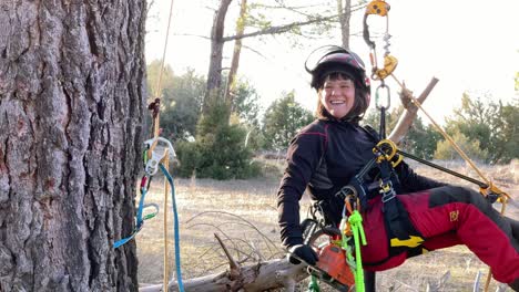 Woman-smiling-hanging-from-a-pine-tree,-attached-with-professional-climbing-equipment-for-gardening,-wearing-a-helmet,-with-a-safety-harness-and-several-carabiners-and-ropes