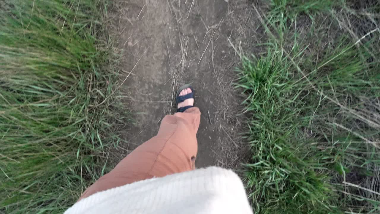 Woman's feet wearing sandals walking forward on a grassy dirt path in first person view