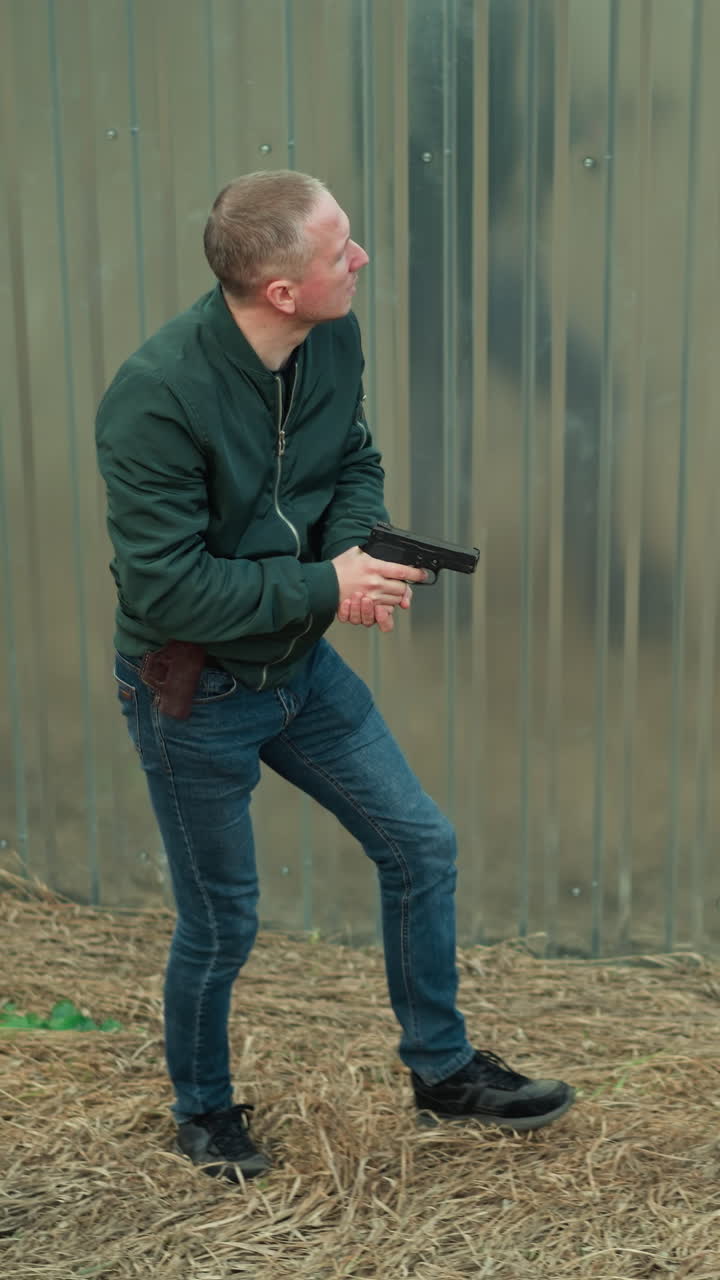 una vista de cerca de un hombre con una chaqueta verde sosteniendo una pistola mientras se mueve con cautela a lo largo de una valla metálica en un área abierta y seca