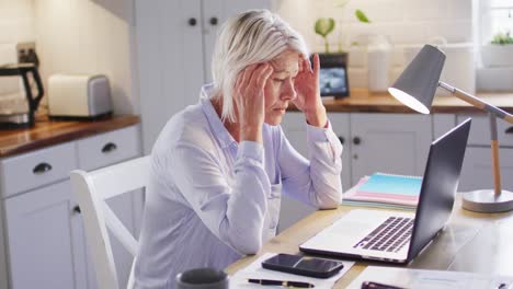 Thoughtful-senior-caucasian-woman-sitting-at-table-in-kitchen,-using-laptop