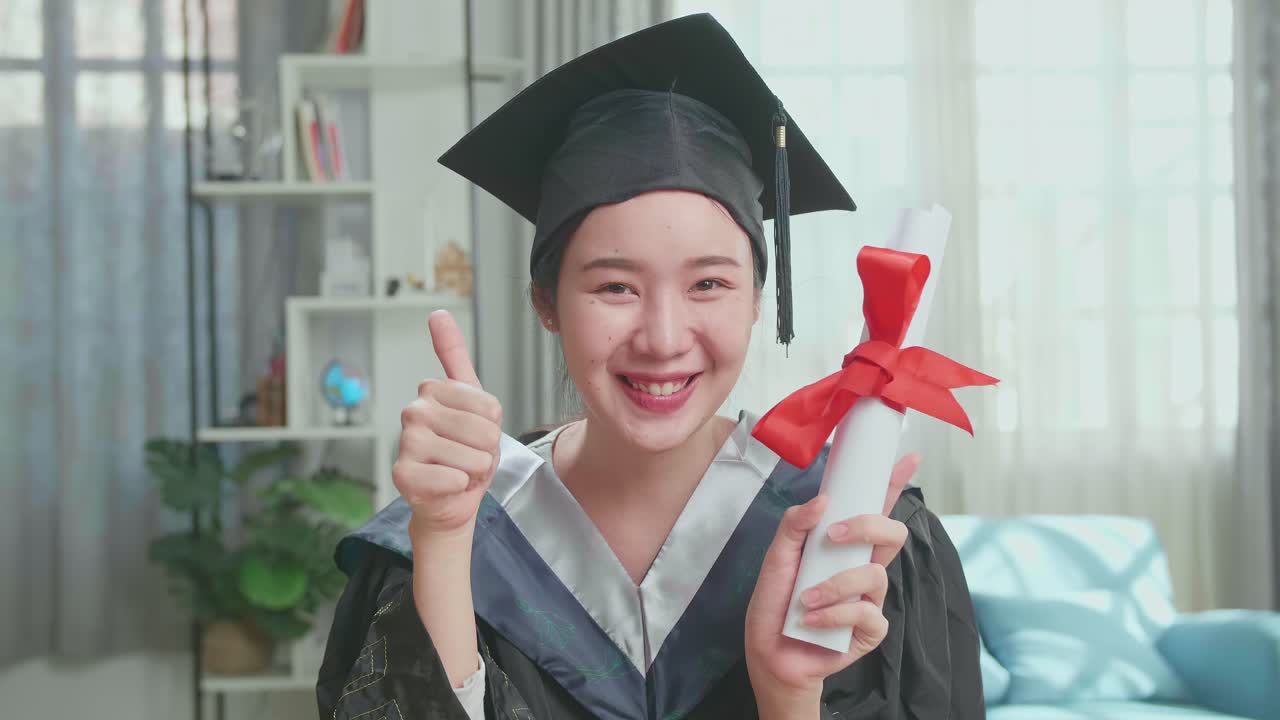 Excited Asian Woman Holding A University Certificate, Smiling And Thumb Up To Camera. Pretty Female Graduate Wearing A Graduation Gown And Cap Sitting On The Living Room