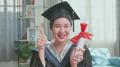 excited asian woman holding a university certificate, smiling and thumb up to camera. pretty female graduate wearing a graduation gown and cap sitting on the living room