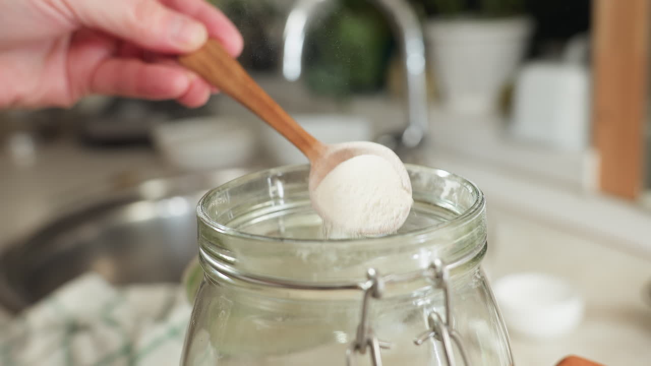 Hand view of chef scooping corn flour with wooden spoon from glass jar, gently shaking to level for accurate measurement, with visible dust particles rising in warm kitchen