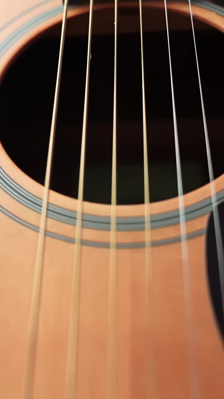 Movimento macro verticale verso l'alto dal ponte della chitarra acustica lungo le corde fino alla buca della risonanza, rivelando la venatura del legno, il dettaglio della rosetta e l'illuminazione calda dello studio per b-roll musicale.