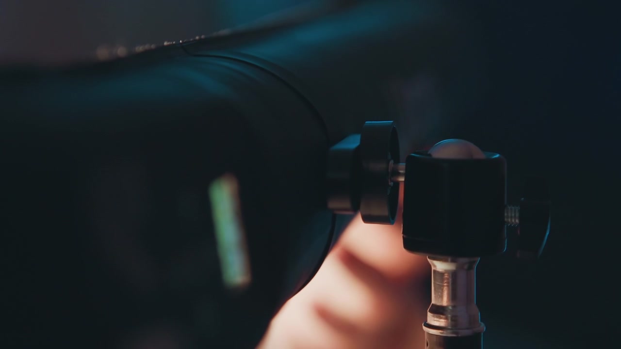 Tight low-angle close-up showing camera connected to tripod head, support post, and locking knob with subtle tilt, revealing recent adjustment, stable mounting, and moody mixed lighting