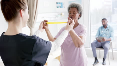 Mujer-Mayor-Diversa-Haciendo-Ejercicio-Con-Banda-Y-Doctora-Asesorando-En-El-Hospital,-Cámara-Lenta