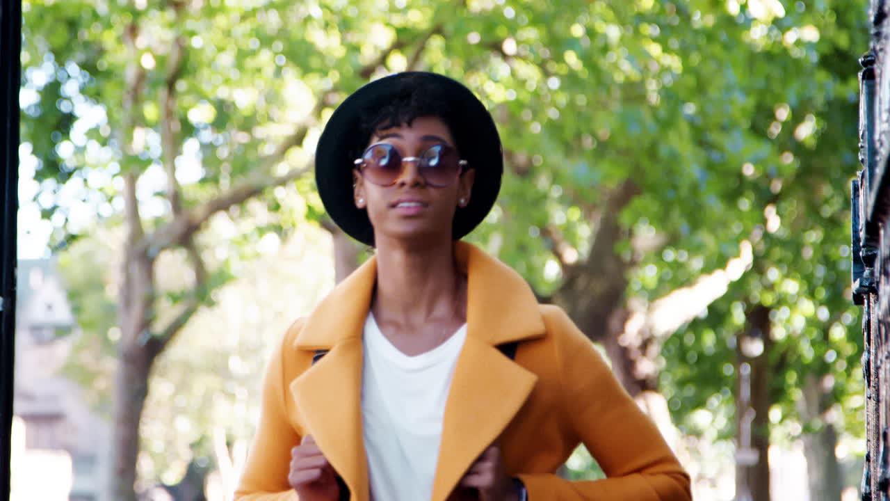 Low angle close up of a fashionable young black woman wearing a hat, sunglasses and a yellow coat walking in the street, head and shoulders