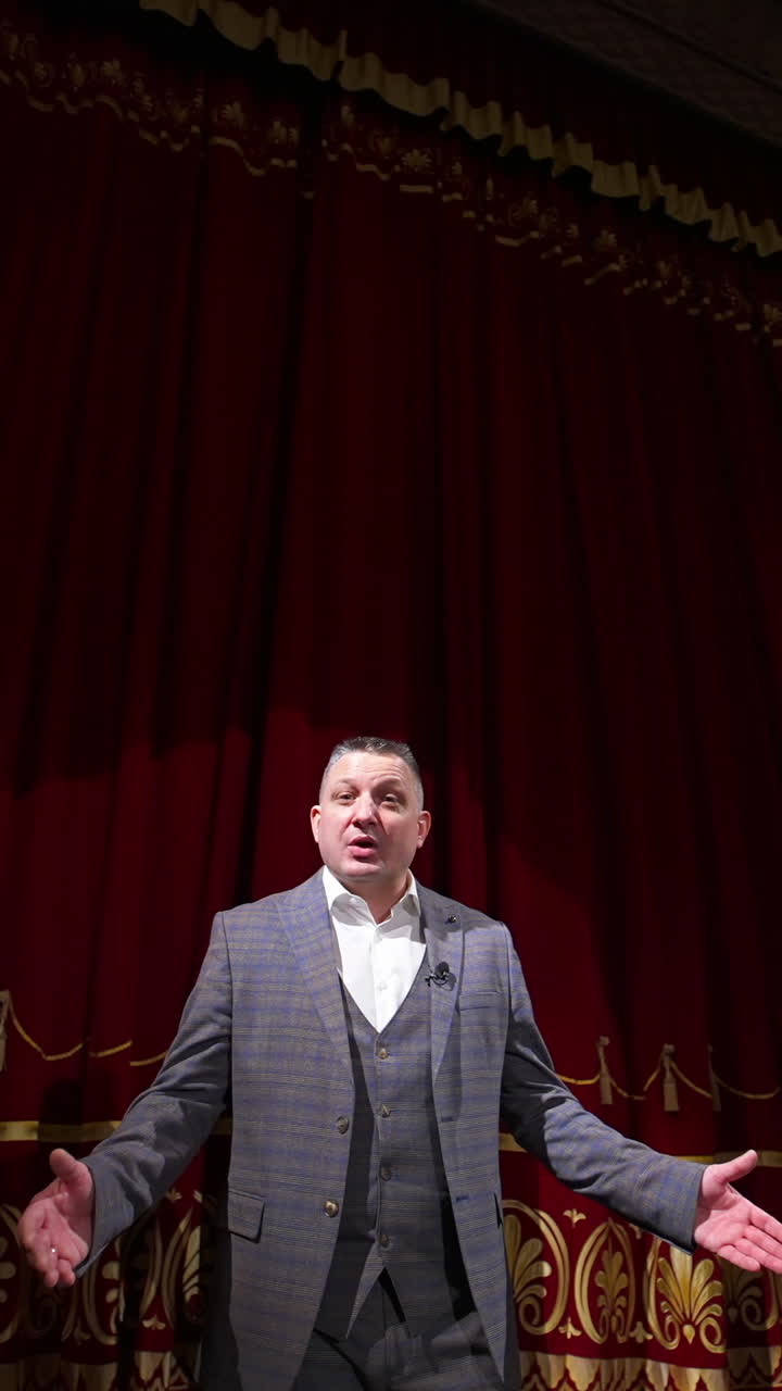 Theater host on red curtains background. Man in grey costume talking on a big stage. Portrait of an actor performing on stage. Vertical video