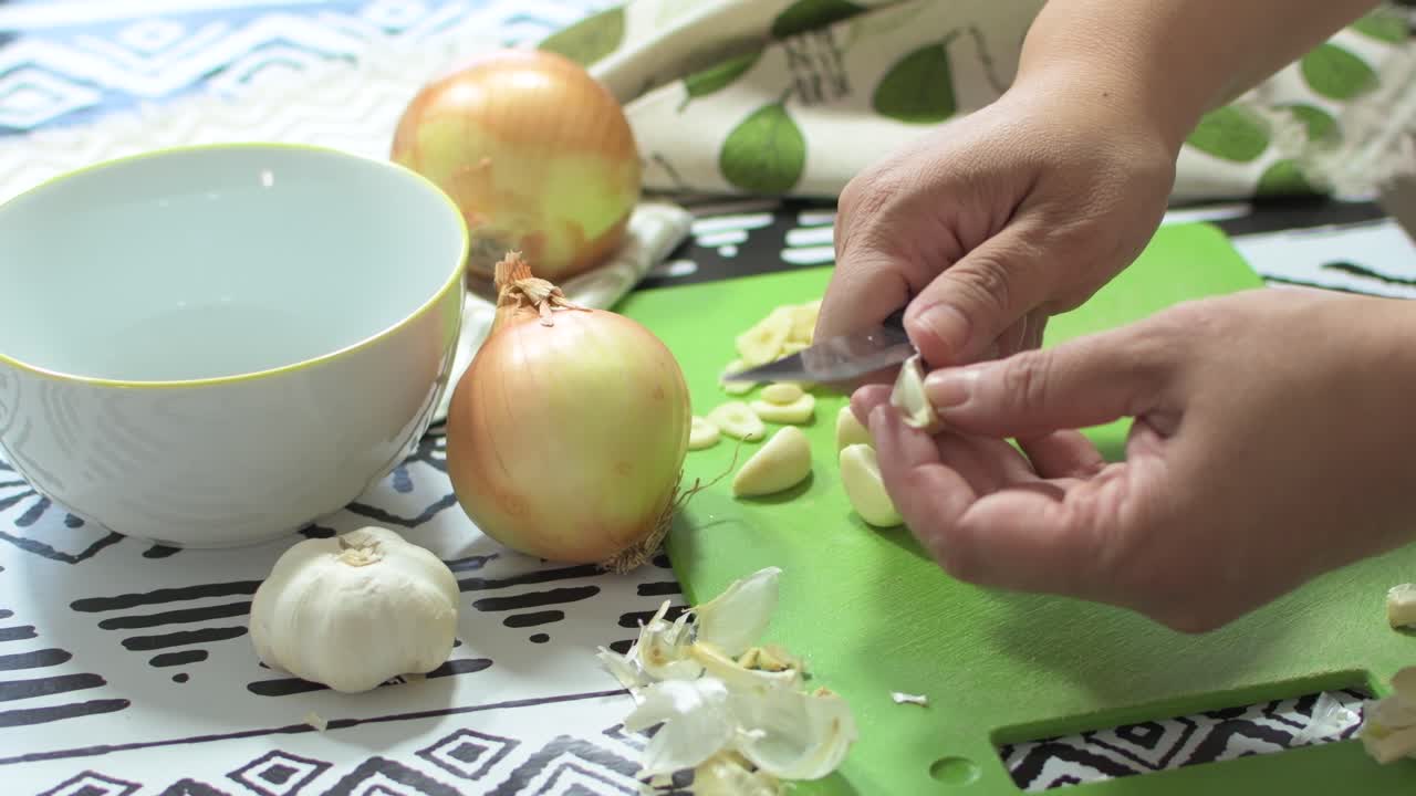 Woman's hands peeling garlic, on a green cutting board, next to onions and a white bowl, on a kitchen table.