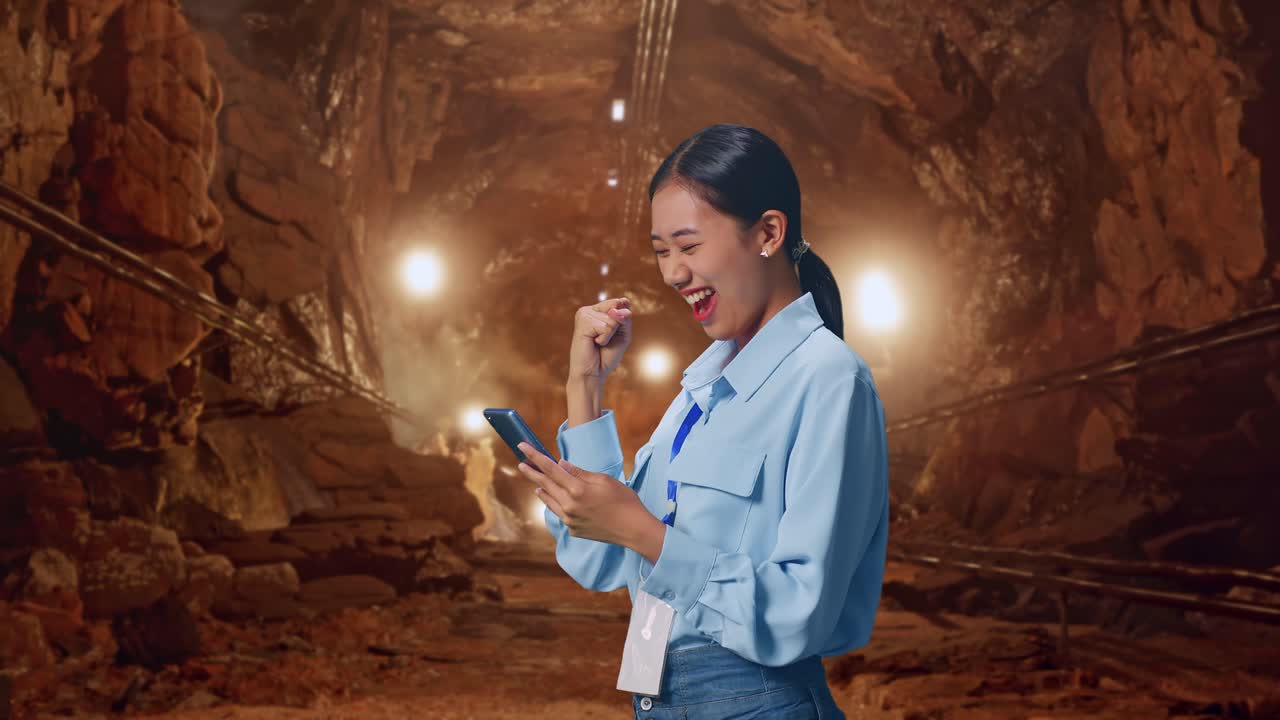 Side View Of Asian Female Professional Worker With Her Smartphone In Underground Mine Tunnel, She Raises Her Fist Up With Screaming Goal After Check On The Smartphone