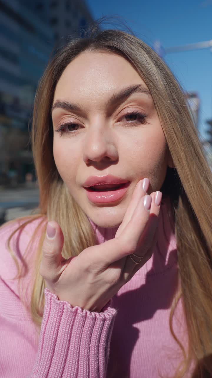 Vertical close-up - blondy young woman touching up her lip makeup at city street looking directly at camera smiling