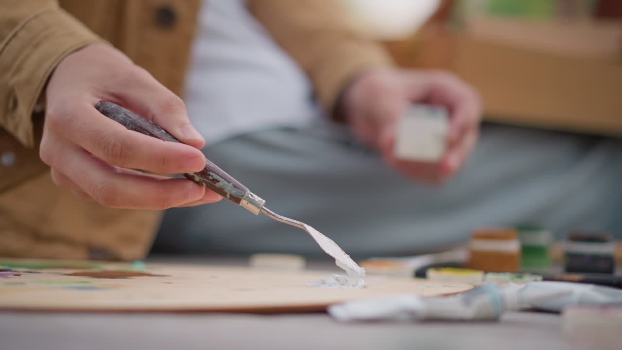artist preparing white paint with knife, closeup of hands squeezing paste from small container, palette receiving thick white mixture, jars of color lined on bench, casual jacket and jeans, deliberate