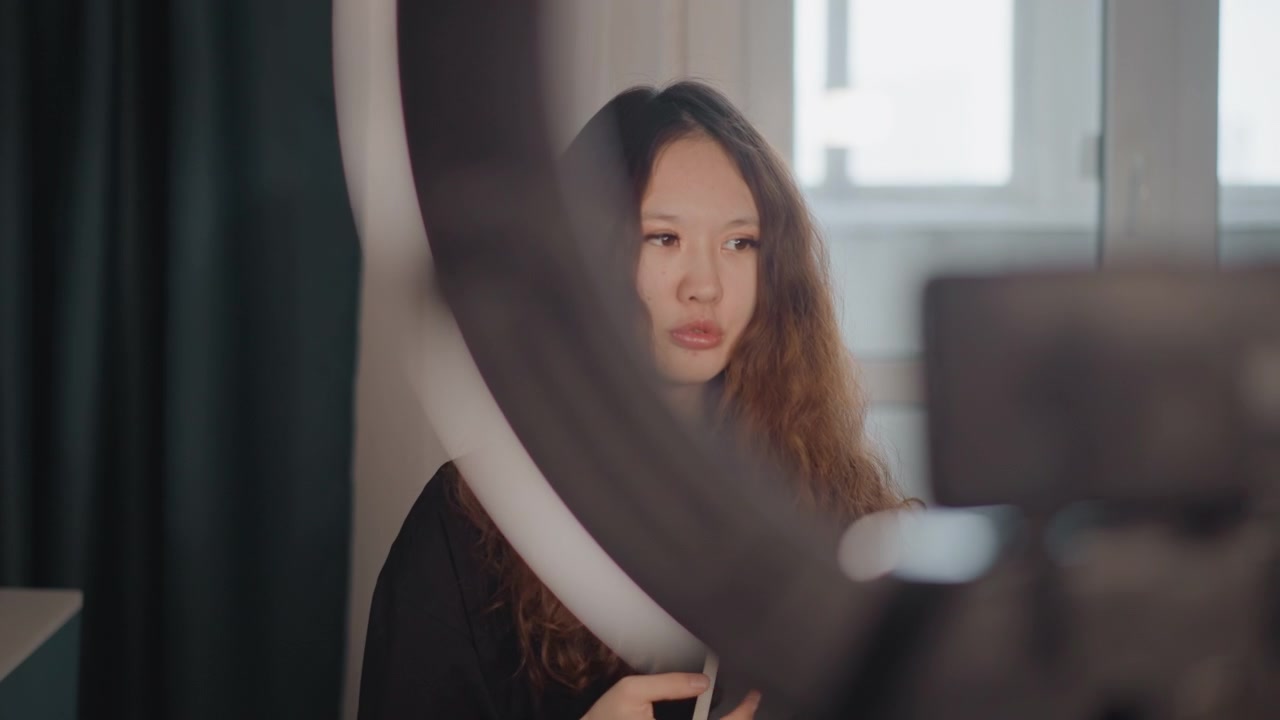 Giovane donna seduta in un interno domestico moderno controlla lo smartphone accanto a una ring light luminosa e al riflesso dello specchio, con un'espressione concentrata, luce diurna soffusa, un'atmosfera da ritratto calma e una sottile routine di cura di sé