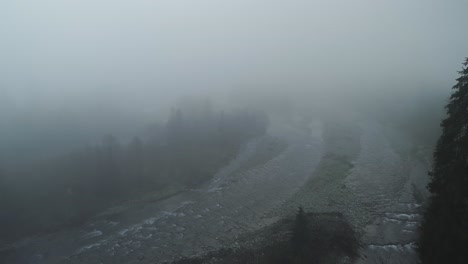 Paisaje-De-Terror-Con-Niebla-Espesa-Y-árboles-Oscuros-Junto-Al-Río-Rocoso,-Aéreo