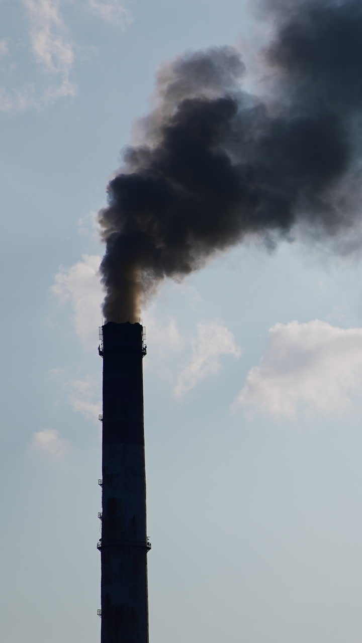 Two factory chimney pipes producing heavy dark smoke. Industry polluting the atmosphere. Cloudy sky backdrop. Vertical video