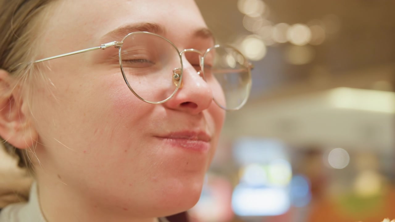 Bloguera gastronómica sonriendo mientras prueba una hamburguesa, retrato de primer plano con gafas y bokeh suave, rostro expresivo mostrando anticipación y luego deleite, ambiente cálido de restaurante interior e iluminación tenue