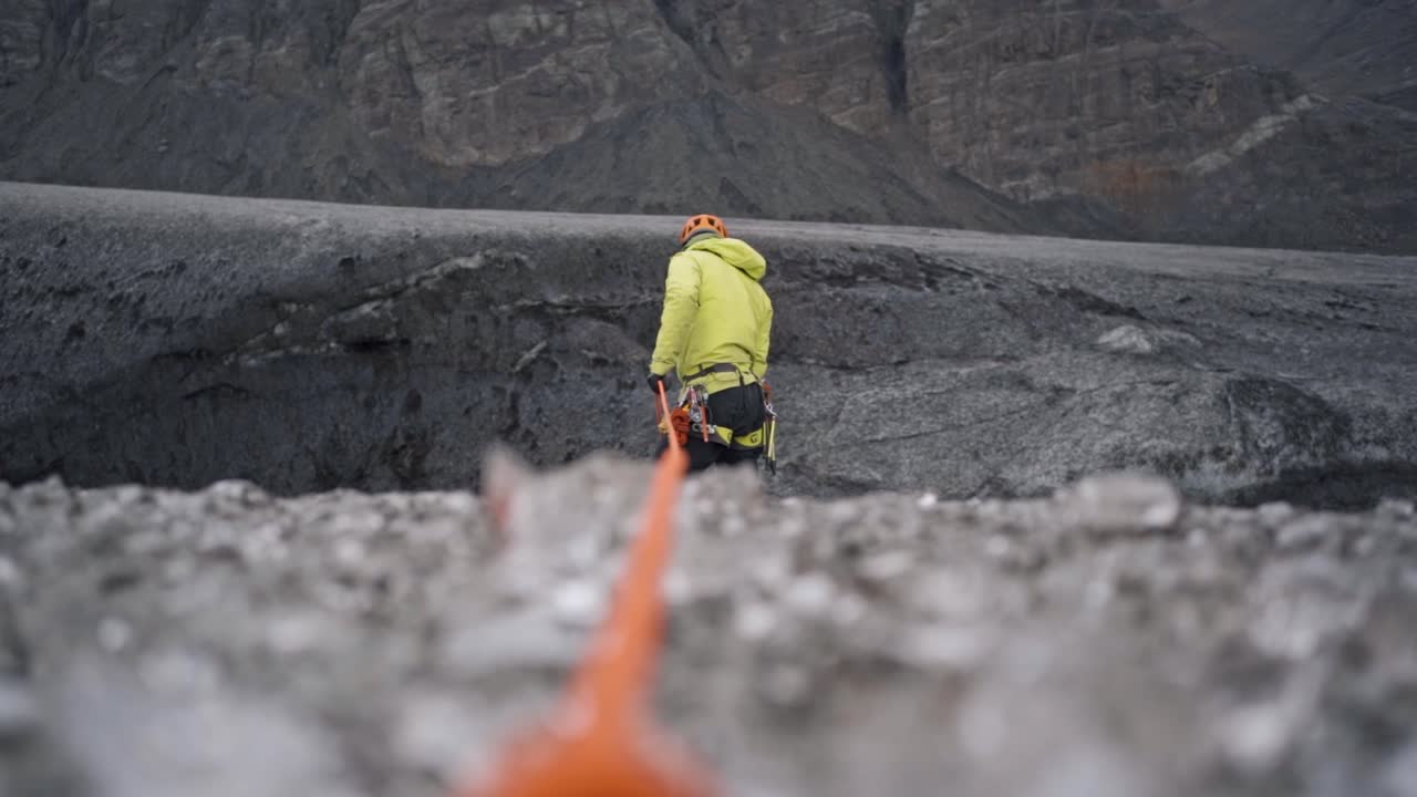 A dynamic shot features a person in bright yellow outdoor gear, seemingly on a rope, positioned against a dark, rugged, and possibly icy or rocky glacial landscape